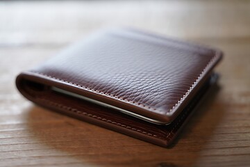A Rich Brown Leather Wallet Closed on a Light Brown Wooden Surface Detailed Closeup View Showing Stitching and Texture