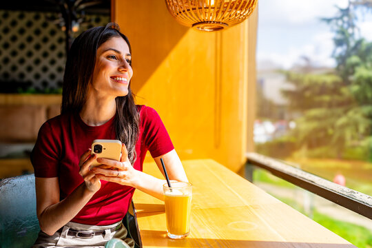 Young woman enjoying a fruit juice in a cafe while holding her smartphone and looking outside the window