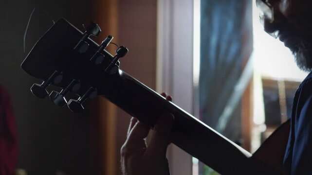 A contemplative and musical mood is captured by a close-up, low-angle view of a man playing a guitar, with the headstock and tuning pegs prominent, subtly lit from behind