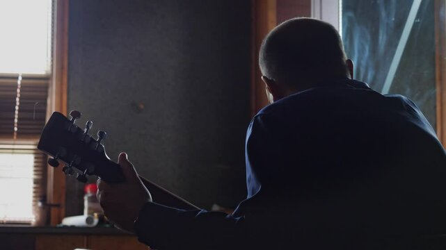 An intimate and dedicated mood is captured by a close-up of a man playing a guitar, with his face obscured by shadow and backlight, highlighting his intense focus on the instrument's strings