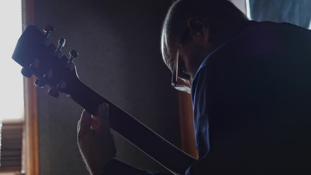 An intimate and dedicated mood is captured by a close-up of a man playing a guitar, with his face obscured by shadow and backlight, highlighting his intense focus on the instrument's strings