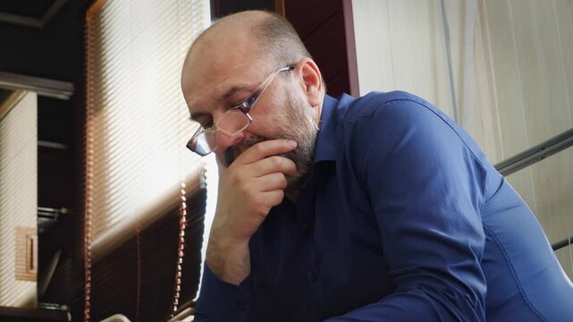 A focused and contemplative mood is captured as a bald man with a beard and glasses sits, intently looking down at his smartphone. This shot is ideal for depicting digital engagement, online research