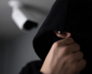 Mysterious figure in black hoodie conceals their identity, partially obscured by mask, while security camera looms in background, capturing tense atmosphere of secrecy and surveillance