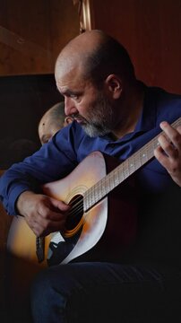 An intimate and dedicated mood is captured as a bald man with a beard intensely plays an acoustic guitar, his gaze fixed on the fretboard. This shot is ideal for depicting musical talent