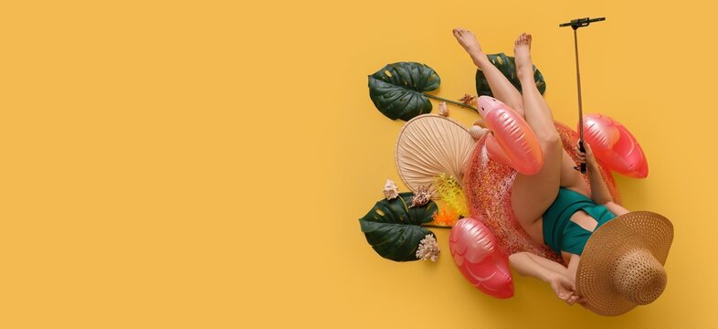 Beautiful young woman in swimsuit,with inflatable ring and tropical leaves taking selfie on yellow background with space for text, top view