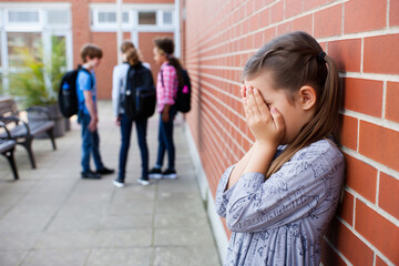 A girl stands and cry against a wall with her face covered, visibly upset while classmates chat nearby in a school courtyard. school bullying concept. Generative AI.