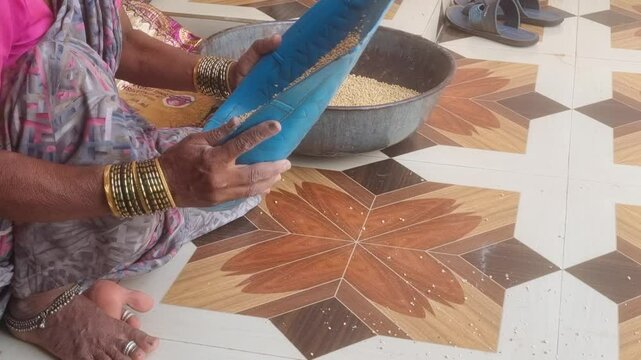 A housewife winnows jowar (sorghum) grains outside her rural home, using a flat basket to separate chaff from grain which is a vital step for clean, healthy food preparation in daily village life.