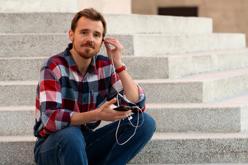 Man seated on steps adjusting earphones connected to smartphone. Guy with beard and modern hairstyle wearing in blue, red and white plaid shirt walking through the city streets in urban background