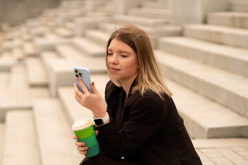Young woman checking phone while holding coffee cup outside. Natural feel with soft light. Casual, confident pose and expression add genuine and approachable vibe