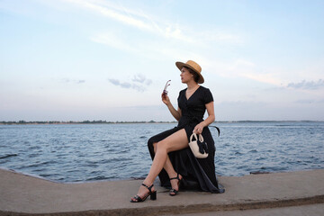 Woman in polka-dot dress gazes at sea holding sunglasses.