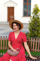 Smiling woman in red wrap dress and hat poses by building.