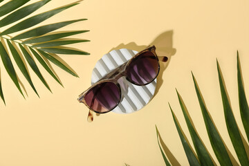 Stand with stylish print sunglasses and palm leaves on pale yellow background