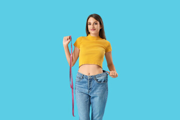 Beautiful young woman in loose jeans with measuring tape on blue background. Weight loss concept