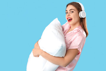 Portrait of pretty young woman in pajamas with headphones and pillow on blue background