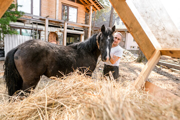 rustic man with  horse in rural village near  forest.  horse farm is surrounded by nature, hay is stacked nearby,  peaceful and traditional.  man is caring for  horse