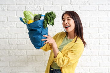 Young Asian woman with blue shopper bag full of products on white brick background