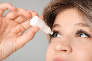 Beautiful young woman using eye drops on grey background, closeup