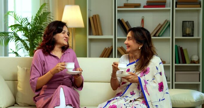 Senior Mother Daughter drinking tea, Indian asian old mother in saree with young daughter sitting together on sofa in modern living room enjoying hot morning beverage while chatting bonding joyfully