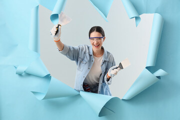 Young woman with putty knives visible through torn blue paper on light background