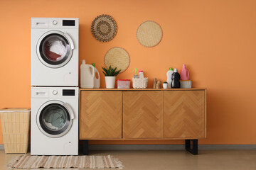Modern washing machines with laundry and chest of drawers with detergent near orange wall in room