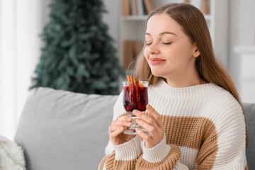 Beautiful young woman in warm sweater with glass cup of hot mulled wine sitting on sofa at home