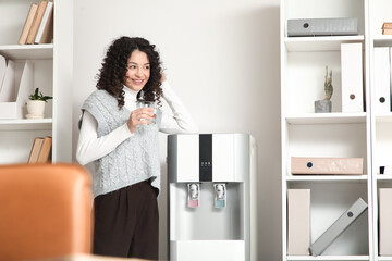 Young businesswoman with glass of water near cooler in office