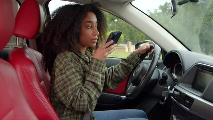 Positive charming black female driver with stylish haircut sitting in vehicle, recording voice message on cellphone while driving car in traffic jam in rush hour.