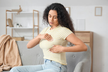Young African-American woman checking her breast on sofa at home. Cancer awareness concept