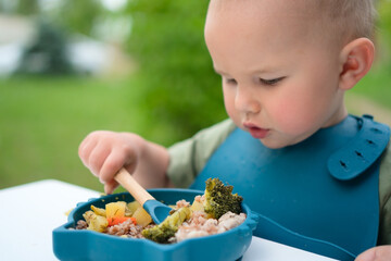 Toddler eating with a spoon from a bowl outdoors. Healthy baby meal with vegetables on a sunny day.