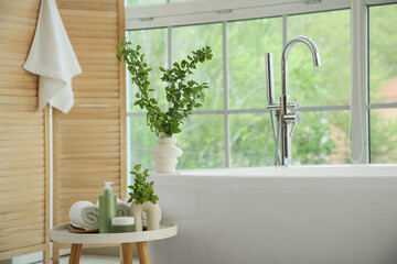 Interior of stylish bathroom with bathtub, folding screen, cosmetic products and green blossoming branches in vases on table near window