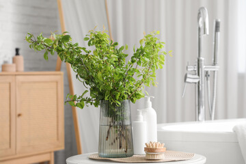 Vase of green blossoming branches with cosmetic products and massage brush on table near bathtub in stylish bathroom