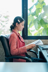 Businesswoman in a red blazer reading papers at a desk in a bright office with a view of greenery outside.