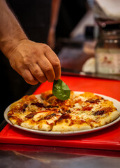 Close-up of a chef placing a fresh basil leaf on a sliced Italian pizza in a traditional restaurant setting, highlighting authentic cuisine and craftsmanship.