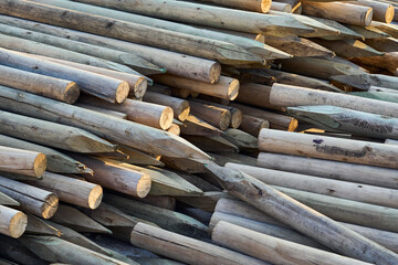 A stack of wooden stakes with sharpened ends, arranged in a pile at a construction or agricultural site. The natural texture and grain of the timber are clearly visible, with warm sunlight casting sof