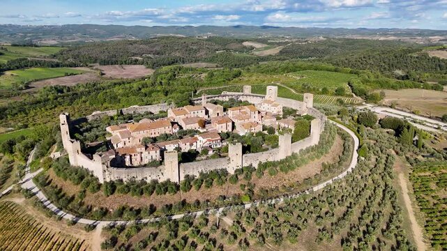 Aerial view of a medieval walled village of Monteriggioni in Tuscany, Italy with towers and red-tiled roofs, surrounded by lush greenery and vineyards.