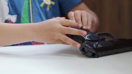 The hands of a young child are seen holding and manipulating a black toy gun, which rests on a white table indoors.