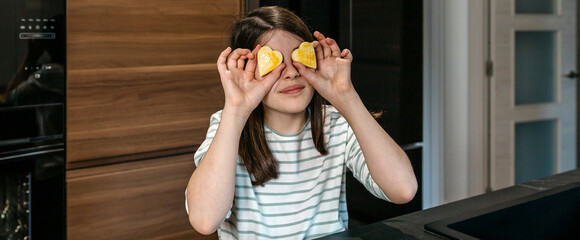 Banner of young girl having fun in the kitchen, holding heart shaped cookies over her eyes, expressing playfulness and enjoyment. Happy little girl looking at camera while covering eyes with biscuits.