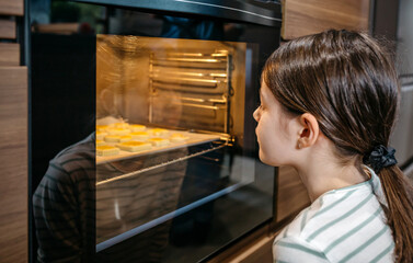 Little girl watching cookies baking in oven, waiting for them to be ready, enjoying the sweet smell and anticipating the delicious taste. Cooking at leisure time concept. Left copy space available.