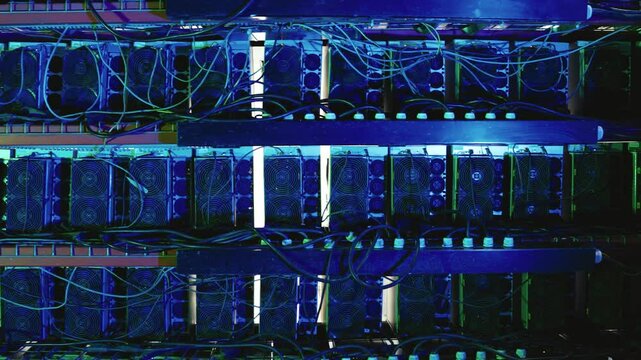Multiple rows of Bitcoin mining rigs with cooling fans and tangled cables, lit by blue light in a data center. Pan right, close up