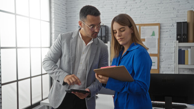 Man and woman collaborating in modern office discussing documents and using digital devices, conveying teamwork in professional work environment.