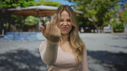 Woman with flowing blonde hair flips middle finger in park under sunlight; defiance rebellion anger...