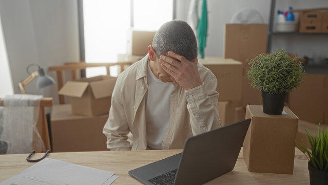 Man sitting stressed in a new home's living room amidst moving boxes, contemplating over a laptop, expresses relocation anxiety and adjustment stress.