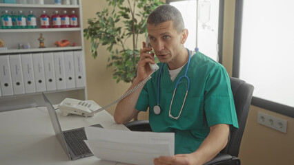 Man wearing green scrubs with stethoscope talks on phone holding papers in a modern clinic office...