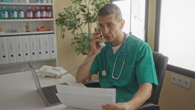 Man wearing green scrubs with stethoscope talks on phone holding papers in a modern clinic office with a laptop and organized shelves in the background. - Powered by Adobe