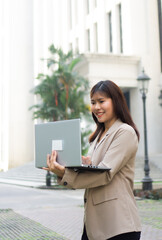 Young happy Asian businesswoman working on laptop in city. Confident female executive using...