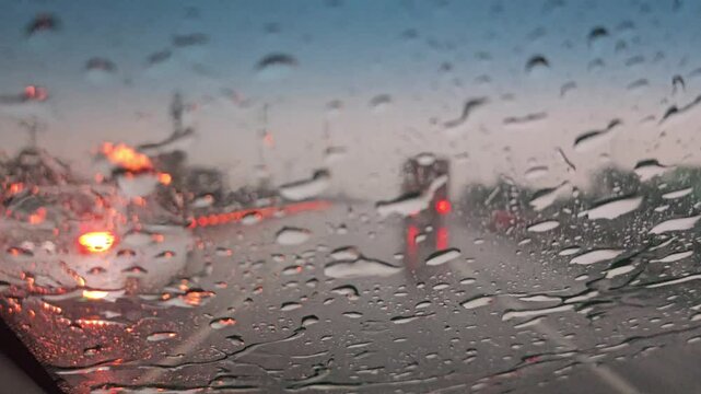 A close-up view through the rain-soaked west glass window of a motor vehicle reveals blurred lights, passing cars, and traffic reflections, as windshield wipers sweep on glass under falling rain drops