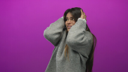 Young hispanic woman covering ears in discomfort, wearing gray sweater against vibrant pink...