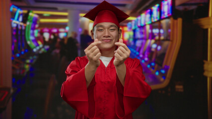 Young man in red graduation gown makes heart symbol with fingers in vibrant casino, surrounded by...