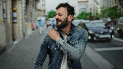 Hispanic man with a beard wearing denim jacket on a busy street, reflecting outdoors, possibly...
