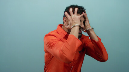Prisoner man wearing orange jumpsuit and handcuffs, gesturing expressively against blue background...